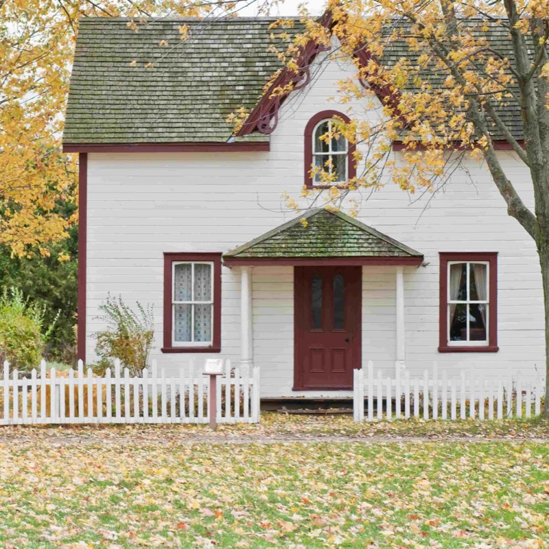 A small white cottage with a picket fence under an autumn tree
