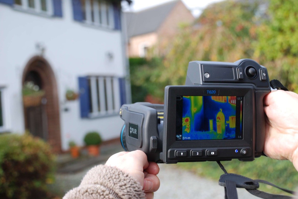 A handheld thermal imaging camera showing a color heat map of a house
